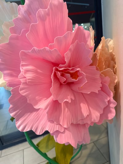 This media file displays a close-up of a large, artificial pink flower with ruffled petals. The flower is positioned prominently in the foreground, taking up most of the frame. Behind it, elements of a background suggest an indoor setting, possibly a commercial or decorative space, with hints of a reflective surface and tiled walls. A partially visible light source casts shadows and highlights on the flower's petals, emphasizing their texture and layered structure. The lighting suggests it could be daytime. No people are visible. There is no visible text. The image is cropped to focus on the flower, with a green stem and a hint of another flower or petal in a pale peach hue visible in the upper left corner. The overall impression is of a decorative element, likely part of a larger arrangement.