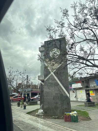 The image depicts a view from inside a vehicle in Coacoatzintla, Mexico, looking towards a town square. A tall, gray stone monument with a clock face and a diagonal white cross adorns the square, partially obscured by tree branches. Buildings and businesses surround the square, with parked cars and pedestrians visible. Produce crates sit near the monument. The sky is overcast.