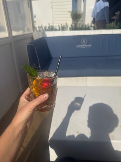 The image captures a first-person perspective of a hand holding a cocktail glass, with a shadow cast on a bright surface. The drink contains ice, a red fruit (possibly a cherry or strawberry), and a mint leaf garnish. A black straw is visible in the glass.

In the background, there is an outdoor setting, featuring a blue cushioned bench with a brand logo. Through a window, one can discern greenery and a building. A partial view of a person in a light-colored shirt is visible on the bench.

Based on the presence of the brand logo on the bench and contextual knowledge, the location is identified as the restaurant Coast Knokke-Heist in Knokke-Heist, Belgium.