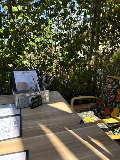 Here's a detailed description of the image:
**Scene:** The photo captures an outdoor dining setting, likely at a restaurant or cafe. A wooden table takes up a significant portion of the frame, with a colorful chair visible at the top. The table is set with menus and a utensil holder containing cutlery. A sign indicates a reservation for "Karen x3" at 13:00. Lush green foliage fills the background, creating a garden-like ambiance.
**Subjects and Objects:**
* **Table:** A wooden table with a light, natural finish.
* **Chair:** A chair with a colorful patterned seat.
* **Menus:** Multiple menus are placed on the table.
* **Utensil Holder:** A metal holder containing forks, knives, and spoons.
* **Reservation Sign:** A small sign indicating a reservation for "Karen x3" at 13:00.
* **Foliage:** Dense green foliage forms a backdrop to the scene.
**Environment:** The setting appears to be outdoors, possibly in a garden or patio area. The presence of foliage suggests a natural environment.
**Location (Based on OCR):** West Oxfordshire, United Kingdom (due to the mention of "Cherry Tree Cablak" on the menu, which could be a local specialty or dish).