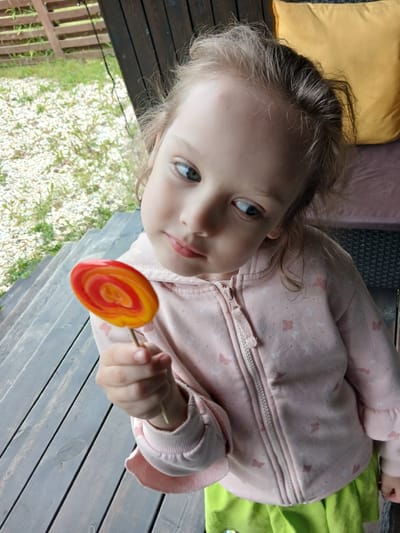 A young girl with blonde hair is holding a swirl lollipop with red and yellow colors. She is wearing a pink hooded jacket with butterfly patterns and a green skirt. The girl is standing on a wooden deck with wooden railings and green grass visible in the background. A yellow pillow is also visible in the background. The scene appears to be outdoors, possibly on a porch or deck in Verkhneye Dubrovo, Russia.