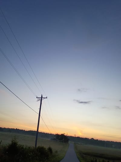 The image depicts an outdoor scene at dusk. A telephone pole with wires stretches diagonally across the frame, from the lower left to the upper right. The pole is silhouetted against a clear sky that transitions from a deep blue at the top to lighter shades of blue and orange near the horizon. A paved road curves into the distance, flanked by grassy fields and a line of trees. The lighting suggests it is either sunrise or sunset. There are no people or animals visible in the frame. The location is identified as being in Jeffersonville, United States.