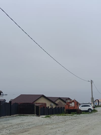 The image depicts a street scene in Zarubino, Russia, under an overcast sky. In the foreground, a gravel road leads towards several houses. A dark, vertical fence runs along the left side of the road, separating it from the properties. Behind the fence, a row of houses with reddish-brown tiled roofs is visible. One house has beige brick siding, and another has darker wood siding. An air conditioning unit is mounted on the wall of the brick house.
To the right of the houses, a prominent orange excavator is parked on a grassy area. Beside the excavator, a white SUV is parked. A utility pole with a street light stands to the right of the vehicles. In the background, a body of water, likely the sea, is visible under a thick fog or haze. Several rock formations are protruding from the water in the distance. Another house with a dark roof is visible further back on the left. The overall impression is of a coastal settlement on a cloudy day.