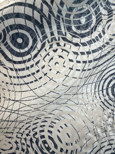 This media file is a close-up, top-down view of a fabric with a black and white abstract pattern. The pattern consists of concentric circles, some filled with solid black, others with a grid-like design of small squares, and some are outlined with thin black lines. There are also scattered thin black lines that crisscross the fabric, forming a net-like or chaotic scribble effect. The fabric itself appears to be textured, with visible woven threads and some slight creasing or rippling, indicating it is likely a garment or a piece of material. The lighting is even, with no strong shadows, suggesting an indoor setting or diffused natural light. There are no people, objects, or discernible scene components; the sole subject is the patterned fabric. No text is visible in the image. The context provided places this image in Dorsten, Germany, but there are no visual cues within the image to support or contradict this.
