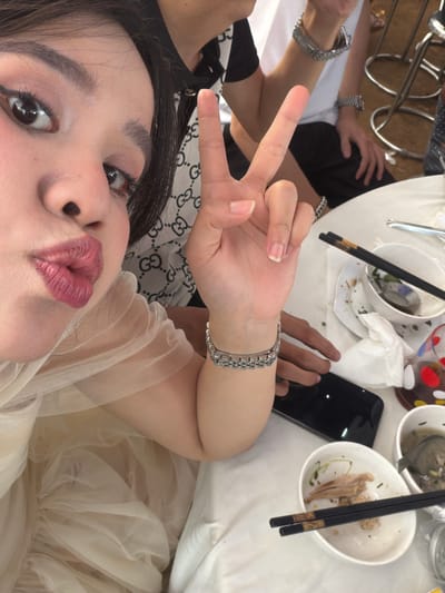 A close-up selfie of a woman with dark hair, fair skin, and pink lipstick, making a peace sign. She is wearing a beige tulle dress. The background includes a table with dirty bowls, chopsticks, a black smartphone, and a person wearing a watch and a black and white patterned garment. The location is not specified.