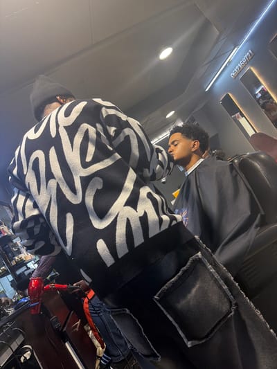 The image, originally rotated 90 degrees clockwise, depicts a scene inside a barbershop or hair salon in Blouberg, South Africa.
**Subjects:**
Two individuals are visible. One, presumed to be the customer, is seated in a black barber chair, seen in profile facing left. This individual has dark curly hair, a light brown skin tone, and is wearing a white collared shirt under a black haircutting cape. Their eyes are cast downwards. Partially visible in the foreground, from the back, is another person, likely the barber or stylist. This individual is wearing a black beanie and a distinctive black and white sweater with an abstract, wavy pattern. They also appear to be wearing dark, distressed jeans with frayed edges and an orange lining.
**Activity or event taking place:**
A hair styling or haircutting session is in progress, with the customer comfortably seated.
**Notable details:**
The interior features dark walls and ceiling, illuminated by several bright recessed spotlights and linear strip lights. One strip light highlights overhead mirrors that reflect parts of the room. A red hairdryer is placed on a dark wooden surface in the lower right. The general ambiance is dimly lit, with focused lighting on the immediate area.
**Visible text:**
On the dark ceiling, illuminated by a strip light, the numbers "0512169721" are displayed.