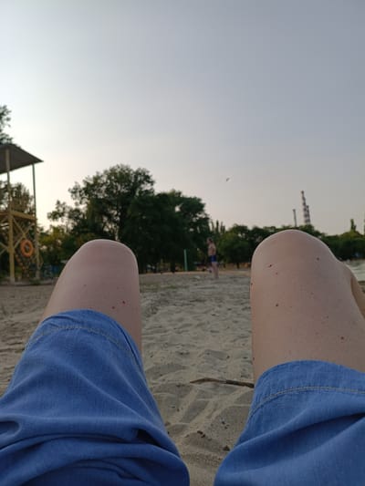 The image shows a low-angle perspective of a person's legs, wearing denim shorts, resting on a sandy beach. The sand is light brown and appears slightly damp in places. In the background, there is a lifeguard tower with an orange life preserver. Several trees are visible, with dense green foliage. Beyond the trees, a body of water is present, and on the far side of the water, industrial structures, including smokestacks, are visible. The sky is a pale, overcast gray. In the mid-ground, a person in blue shorts is standing. The overall scene suggests a relaxed atmosphere at a beach, possibly in the late afternoon or early evening. No specific details about Nezavertaylovka, Moldova, are evident from the image itself.