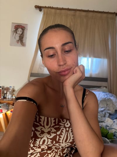 A woman is sitting on a bed, leaning her head on her hand. She is looking down. The room appears to be a bedroom, with a bed, curtains, and a picture on the wall. A table with bottles on it is visible in the background. The woman is wearing a brown and white patterned top.