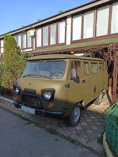 The image shows a parked, olive green UAZ-452 van. The vehicle is parked on a brick and asphalt surface, adjacent to a building with a multi-windowed facade, likely a commercial or residential structure. The building's design features several windows with white curtains and brown frames. A small section of green metal fencing is visible to the right of the van. The license plate is visible on the front bumper and indicates the location as Рыбхоз, Russia. The scene is captured during daylight under a clear sky. No people are visible in the frame.
