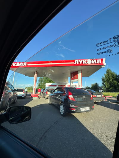 This image captures a scene at a Lukoil gas station, likely in Kirovo-Chepetsk, Russia, on a clear, sunny day. The perspective is from inside a vehicle, looking out through the side window and windshield. A line of cars is waiting to refuel, indicating a busy period at the station.
The dominant feature is the red and white canopy of the Lukoil gas station, with its prominent logo visible. Beneath the canopy are several gas pumps, with at least two cars actively refueling. In the foreground, a black Hyundai Accent is parked, facing away from the camera. Its license plate is partially visible, reading "P091OE 123". Other vehicles, including SUVs and sedans, are also in the queue.
Several individuals are present. One person, wearing a light blue shirt, is standing near a pump, possibly attending to a vehicle. Another individual, dressed in a red jacket and shorts, is seen near the pumps, appearing to be involved in the refueling process. The surrounding environment includes asphalt pavement, green trees in the background, and a clear blue sky overhead. Reflections are visible on the car window and the gas station canopy, suggesting a bright day. The overall impression is one of a typical, albeit busy, day at a Russian gas station.