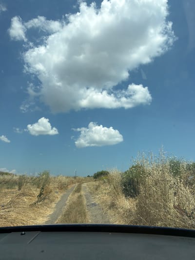 The image displays a roadside view with a dirt path winding through dry, golden vegetation on the right side. The vegetation appears to be tall grass or hay, suggesting a rural or agricultural setting. A thin, dark line runs vertically along the far right edge of the frame, possibly indicating the edge of a vehicle's window or frame, implying the photo was taken from inside a moving vehicle. The sky above is a clear, bright blue, dotted with several fluffy white cumulus clouds. The lighting suggests it is daytime, likely late morning or afternoon, given the brightness of the sun and the clear sky. There are no people visible in the image. The scene evokes a feeling of a journey through a sun-drenched, perhaps warm, rural landscape. The location context is Aheloy, Bulgaria, which aligns with the visual cues of open, possibly dry terrain and a clear sky.