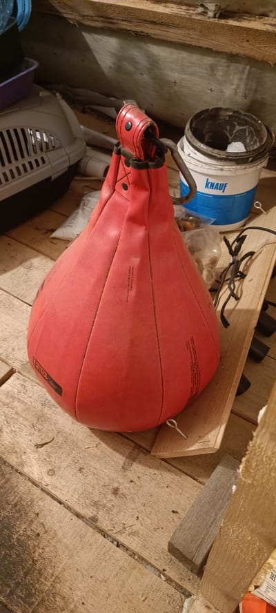 A red punching bag hangs on a metal hook, suspended over a wooden floor. The bag is a pear shape, with stitching lines running vertically up its sides. A black label reading "PRO Class" is visible on the lower left side of the bag. To the right of the punching bag is a white bucket with blue lettering that reads "KNAUF". The bucket appears to contain a white substance. Behind the punching bag, a light-colored cat carrier is visible. The background consists of unfinished wooden planks, suggesting an attic or unfinished room. The lighting is dim, casting shadows across the scene. The overall environment suggests a storage area or workshop. The location is identified as a dacha (country house) in the village of Smorodinka, Russia.