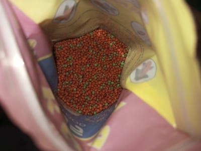 An open bag reveals a dense pile of fish food pellets. The pellets are primarily reddish-brown with scattered green ones interspersed throughout. The bag's interior has a yellow and pink design with small frog illustrations. The bag material appears to be plastic or a similar flexible packaging material.