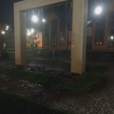 The image depicts an outdoor scene at night, likely in a park or recreational area in Sosnovka, Kyrgyzstan. A large, light-colored concrete archway stands in the foreground, framing the view of several small wooden structures with pitched roofs, resembling cabins or gazebos. These structures are illuminated by bright lights, creating strong reflections and flares. Transparent vertical elements, possibly curtains or decorations, hang within the archway. The ground in front of the archway is wet and appears to be a mix of soil and paving stones, with patches of grass. In the background, trees are visible, also illuminated by artificial lights. The overall impression is one of an outdoor gathering or entertainment space at night, with a wet ground suggesting recent rain or a sprinkler system.
