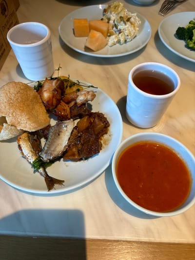 The image shows a table with a meal. On the table are two white cups, one containing clear liquid and the other filled with tea. There is a white plate with various food items, including what appears to be roasted meat, fish, and a large cracker or crisp. Another small white bowl holds a red soup. In the background, there are two more plates with food: one with pieces of melon and a salad, and another with broccoli. The table is a light-colored marble surface. A wooden object with the "Doubletree" logo is visible in the upper left corner, suggesting the location is a Doubletree hotel. The scene is captured from a slightly elevated angle, looking down at the table. There are no people visible in the image. The lighting suggests an indoor setting, likely a dining area. The image does not provide any information to determine the city of Yingzhou, China.