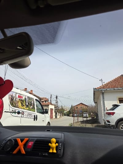 The image, captured from inside a vehicle, presents a static view of a residential street in Gevgelija, North Macedonia. The vehicle's dashboard is visible at the bottom of the frame, featuring an orange 'X' and a small yellow humanoid figurine attached to the central air vents. A rearview mirror hangs from the top of the windshield, from which a red heart-shaped ornament is suspended.

Outside the vehicle, a white commercial van, decorated with large, colorful pictorial decals and Cyrillic text, is positioned on the left side of a paved street. To the right, a light-colored residential building with an orange-tiled roof partially obstructs the view of a parked white SUV. Further down the street, additional single and multi-story buildings with similar tiled roofs are visible amidst sparse trees and bushes. Numerous overhead utility lines crisscross the scene, supported by utility poles. A lone individual is vaguely discernible in the midground, standing near the white SUV on the right. The background consists of a light blue, hazy sky with distant, indistinct hills or mountains on the horizon.