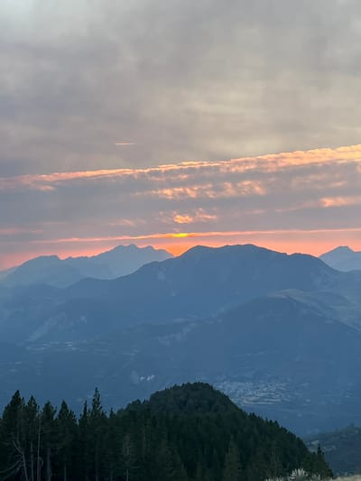 This image depicts a panoramic mountain landscape at sunset, with layers of mountains receding into the distance. The sky is filled with soft, colored clouds, ranging from pale gray to warm orange and pink hues, indicating the sun is low on the horizon. A thin, bright line of sunlight is visible just above the highest mountain range, suggesting the sun is either setting or rising behind it. In the foreground, a dark, densely forested slope with pine trees is prominent. Below the closest mountain range, a distant village or town can be faintly seen nestled in the valley. The overall scene is tranquil and evokes a sense of natural beauty. The location is identified as the municipality of San Juan de Plan/San Chuan de Plan, located in the province of Huesca, Aragon, Spain. The image does not contain any people, animals, or discernible actions.