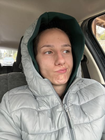 A light-skinned woman with brown hair is lying in the backseat of a car, wearing a light gray puffer jacket with a dark green hood. The label "PULL&BEAR" is visible on the jacket. The car's interior is visible, including the ceiling, a safety handle, and the car seat. Through the car window, blurry objects suggest a natural environment outside with what appears to be a wooden structure with a window. The scene appears to be Hrishteni, Bulgaria, as per the prompt.