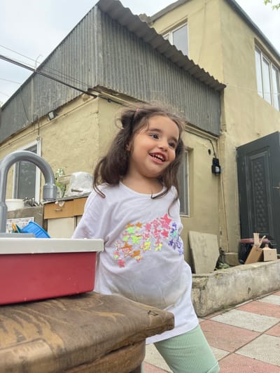 A young girl with long, dark, wavy hair stands in front of a weathered yellow building with a corrugated metal roof. She is wearing a white t-shirt with colorful sequin decorations of various sea creatures and light green ribbed leggings. She is smiling and looking to her right. In the foreground, a red toy sink with a gray faucet sits on a wooden surface. The ground is paved with pink and white checkered tiles. The building appears to be in a residential area with some utility boxes and wires visible on the exterior wall. The overall environment suggests an outdoor setting in a slightly overcast day.