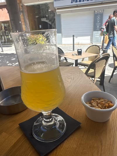 The image captures an outdoor scene on a bright day in Cambrils, Spain. A glass of beer, partially filled, sits on a black napkin on a wooden table. Next to it is a small white bowl filled with peanuts. In the blurred background, there are tables and chairs of an outdoor cafe, a storefront with the name "Mimi," and pedestrians walking by.