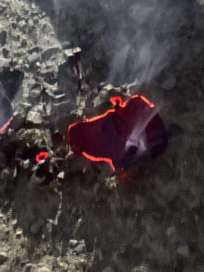 This media file depicts a close-up, aerial view of what appears to be lava flowing or cooling on a dark, rocky surface. The lava is a deep crimson color with glowing orange edges, suggesting intense heat. Wisps of white smoke or steam rise from the lava, indicating ongoing activity or recent cooling. The surrounding terrain is rough and textured, composed of smaller rocks and debris.

The scene is characterized by the stark contrast between the dark, natural landscape and the bright, incandescent lava. The way the lava has spread suggests it is a relatively recent event. There are no visible people or man-made objects, placing the focus squarely on the geological phenomenon. The lighting appears to be natural, possibly daylight, given the illumination of the scene, but the specific time of day is not definitively ascertainable. The weather is not apparent from the image.

The overall impression is one of raw, natural power and elemental forces at work. The image captures a moment in time where the earth's internal heat is manifest on its surface. The location context of Khazar Raion, Azerbaijan, could suggest this is part of volcanic activity or a similar geological event in that region. No text is visible in the image.