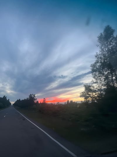 The image depicts a dramatic sunset over a rural landscape. The sky is filled with elongated clouds painted in hues of deep grey, soft blues, and vibrant oranges and reds near the horizon, indicating the sun is low in the sky. The warm colors suggest it is either sunrise or sunset, likely sunset given the deepening shadows. A paved road, marked with a white line, recedes into the distance, curving slightly to the right. Trees and dense foliage line the edge of the road, creating a dark silhouette against the colorful sky. The overall mood is serene and awe-inspiring, with the natural beauty of the sunset dominating the scene. There is no visible text in the image. The location context is Серебрянский, Russia.