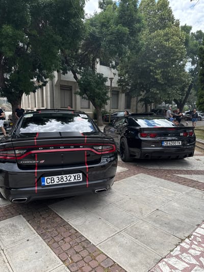 The image shows two black muscle cars, a Dodge Charger and a Chevrolet Camaro, parked on a paved area with trees and a building in the background. The Dodge Charger, in the foreground, has red racing stripes. Its license plate reads "CB 3832 XB". The Chevrolet Camaro, to its right, also has red accents and a spoiler. Its license plate reads "CB 6200 TK". Several people are visible in the background, standing around the cars and conversing. The environment suggests an outdoor gathering or car show. The scene is located in Hadzhidimovo, Bulgaria, as indicated by the license plates and the context provided. The lighting suggests it is daytime.