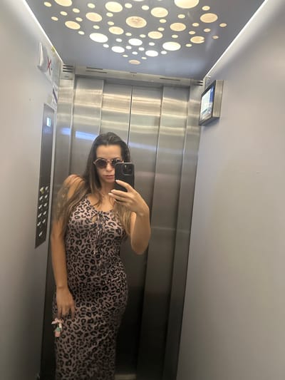 A woman is taking a selfie in an elevator. She is wearing a leopard print dress and sunglasses. She is holding her phone in her right hand, and a keychain with pink and blue charms in her left hand. The elevator has stainless steel walls and a metallic ceiling with circular lights. A "no smoking" sign is visible on the left wall, and a digital screen is mounted on the right wall. The image appears to have been taken in an indoor public space. There is no indication that this location is in Torremolinos, Spain.