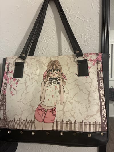 The image displays a tote bag with a graphic print of a young woman holding a camera, with the Eiffel Tower and cherry blossoms visible in the background. The bag is black and beige and has black leather straps. It is hanging on a white, textured wall. The context for the image is a residential interior in Balch Springs, United States of America.