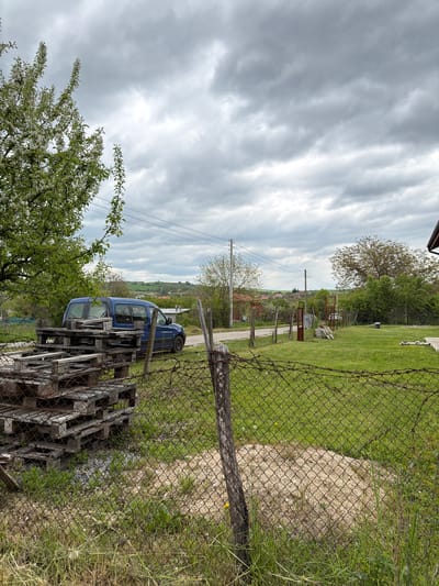The image depicts a rural scene, likely in Dulevo, Bulgaria, although this cannot be definitively confirmed from visual data alone. The foreground features a stack of wooden pallets and a blooming tree with white flowers. A blue car is parked partially behind the pallets. A rusty chain-link fence separates the foreground from a grassy area. Beyond the fence, a dirt road leads toward the background, where several buildings and trees are visible. Power lines stretch across the sky. The sky is overcast with gray clouds.