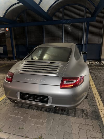 A silver Porsche Carrera 4S is parked on a paved surface with yellow lines marking the parking spot. The car is positioned near a building with a blue and grey facade. The vehicle's rear end is prominently visible, showcasing the taillights, the "Carrera 4S" badge, and the exhaust pipes. The paved surface is cracked in some areas, and there are traces of dirt or water. There's also a tunnel or shelter-like structure near the vehicle. Based on the "ares" sign on the wall in the background, this appears to be near the Ares Parking garage in Wetzikon, Zurich, Switzerland.