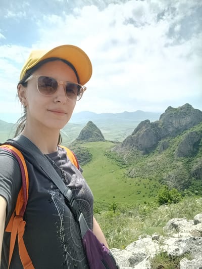 A woman in a yellow baseball cap and sunglasses takes a selfie against a backdrop of rolling green hills and rocky mountains, likely in the vicinity of Hüseynbəyli, Azerbaijan. She wears a grey t-shirt, an orange and purple backpack, and a silver necklace with a charm. The landscape features a distinctive conical hill in the distance, as well as other mountains and rocky formations. The sky is partly cloudy.