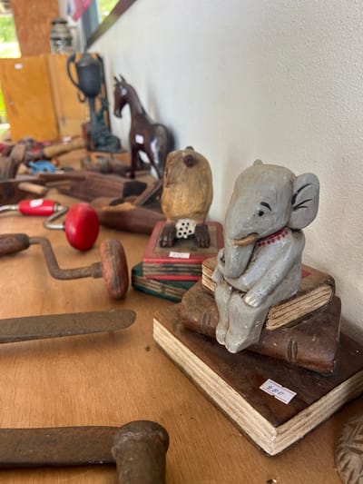 This image shows a collection of handcrafted wooden items and tools displayed on a wooden surface, likely in a market or workshop. The primary subjects are various carved wooden figures and vintage tools, including what appears to be an accordion-playing figure and a carved animal. A price tag with "200" written on it is visible on a wooden base. Among the tools are what look like hand drills and chisels. The setting appears to be indoors, possibly a market stall, given the arrangement of items and the hint of a white ceiling. The lighting suggests it is daytime. The overall mood is one of rustic charm and a display of artisanal craftsmanship.