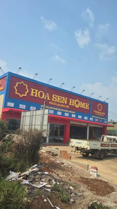 The image shows a commercial building in Trung Hồ, Vietnam. It is a "Hoa Sen Home" store, a chain specializing in building materials and home furnishings. The building is painted with a blue and red color scheme and is decorated with advertisements for various brands. Debris and vegetation can be seen in the foreground. Several vehicles can be seen. There is a kilometer marker beside a road reading Km 169 KRÔNG NĂNG QL.