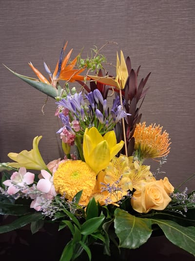 This media file features a close-up, eye-level shot of a vibrant floral arrangement. The arrangement is composed of a variety of flowers and foliage in shades of yellow, orange, purple, and pink, set against a textured, muted brown background that appears to be a wall.
Prominent subjects include a bright orange Bird of Paradise flower, yellow roses, yellow lilies, a yellow chrysanthemum, purple agapanthus flowers, and delicate pink flowers, possibly orchids or carnations. There are also dark reddish-brown leaves and thin, wispy green foliage. One of the yellow flowers in the arrangement is an origami crane.
The lighting suggests it is daytime, possibly indoors with artificial lighting, as there are no strong shadows or indications of natural sunlight. The setting appears to be a well-lit interior space, such as a home, office, or event venue in Ottawa, Canada. There is no activity or event taking place; the focus is solely on the static floral display. No text is visible in the image. The overall impression is one of a carefully composed and colorful decorative arrangement.