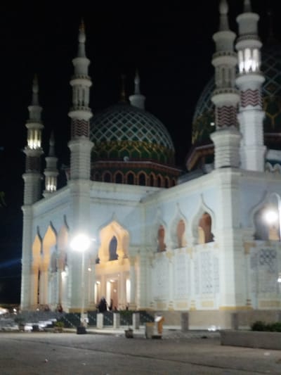 This forensic description details a large, illuminated mosque at night in Kotapantonlabu, Indonesia. The dominant structure is a multi-domed and multi-minaret mosque, predominantly white or light-colored, set against a dark night sky. At least six slender minarets are visible, featuring light-colored shafts; several display reddish-brown chevron patterns on their lower sections and bright internal illumination in their upper sections. Two prominent domes are visible, characterized by intricate green, teal, and gold diamond or square patterns. The mosque's main edifice showcases numerous arched openings, suggesting windows or entrances, some of which are internally lit. Decorative white panels with intricate patterns adorn the lower facade. In the middle-left foreground, a brightly glowing streetlamp illuminates the immediate vicinity. The ground in the foreground is a paved area or roadway, with low planters or barriers visible. Several indistinct human figures are present near the mosque's entrance areas, particularly under the arches on the left side and towards the central entrance, appearing to be standing or moving. The overall scene indicates artificial illumination highlighting the architectural details of the mosque during nighttime.