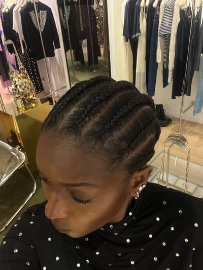 The image depicts an overhead view of a person with dark skin and braided hair (cornrows) looking down and slightly to the left. The individual is wearing a black long-sleeved top adorned with numerous small, reflective studs or rhinestones. In the left ear, a small gold hoop earring and two white stud earrings are visible.

The scene is an indoor retail clothing establishment, likely a boutique, located in Serris, France. The background reveals various clothing items displayed on racks. To the left, a rack holds garments including a black blazer with silver sequin lapels and a black and white polka-dot skirt. A metallic, gold-toned sequined bag rests on a golden surface below. To the right, another clothing rack displays an assortment of garments, including dark blue tops, a white lace-trimmed dress, striped shirts, and denim. A pair of black boots are visible on the floor beneath this rack. Reflective surfaces and mirrors throughout the background show additional garments and reflections of the store interior. The lighting is bright and artificial.