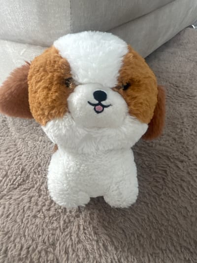 A stuffed animal, appearing to be a dog, lies on a brown, textured surface. The dog is mostly white with brown patches. Its face is white with a black nose and pink cheeks. Part of the dog rests against a gray object, possibly a sofa cushion. The surface underneath the dog appears to be a textured blanket or rug. The location is not discernable from the image.