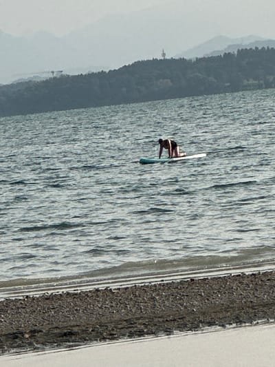 A person is on a paddleboard in the water. The person is in a quadrupedal position, with their knees and hands on the paddleboard. They are wearing dark shorts. The paddleboard is light-colored and appears to be floating on choppy water. In the background, there is a body of water, a treed shoreline, and distant mountains. The image is taken from a low angle, close to the water's edge, which is covered in small rocks. The overall lighting suggests it is daytime. The scene is in Chieming, Germany.