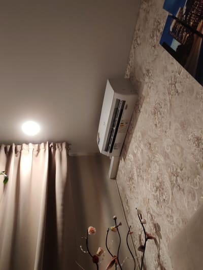 The image shows an indoor scene, likely a room in a residential apartment or hotel. Mounted high on a wall with floral wallpaper is a white air conditioning unit. To the left, a floor-to-ceiling curtain, beige in color, hangs. Above the curtain, a bright, circular light fixture is visible on the ceiling. In the foreground, decorative branches with artificial flowers and beads are arranged, casting shadows on the wall. A portion of a framed artwork depicting a cityscape at night is visible on the upper right side of the wallpapered wall. The scene appears to be in Kotovsk, Russia.