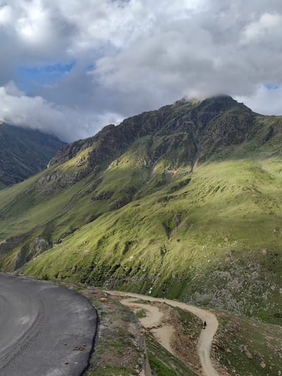 This is a photograph of a winding dirt road traversing a steep, grassy mountain slope in Gramphu, India. The road, characterized by sharp switchbacks, is the central focus of the lower portion of the image, with a section of paved road visible in the foreground. The mountain slopes are covered in vibrant green grass and dotted with rocks, indicating a high-altitude, rugged terrain. Sunlight casts dramatic shadows across the landscape, highlighting the contours of the mountains. The sky is a mixture of heavy gray clouds and patches of blue, suggesting dynamic weather conditions. In the distance, the peaks of the mountains disappear into the clouds. A single figure on a dark-colored motorcycle is visible on the dirt road, adding a sense of scale and activity to the otherwise natural scene. Small, terraced fields are subtly visible on a lower part of the mountain, hinting at human agricultural presence in the area.