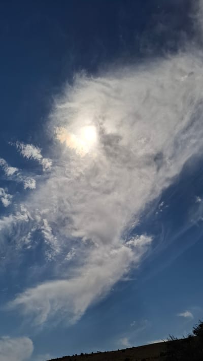 The image depicts a vast, clear blue sky dominating the upper portion of the frame, with scattered white clouds. The sun is visible in the upper center, partially obscured by wispy clouds, casting a bright glow and creating a halo effect. The lower edge of the image shows a dark, hilly terrain, possibly a slope, with sparse vegetation and some small bushes or trees visible. The overall scene is natural and serene, with no people or specific man-made structures clearly identifiable beyond the landscape itself. The location is specified as Boraldai, Kazakhstan.