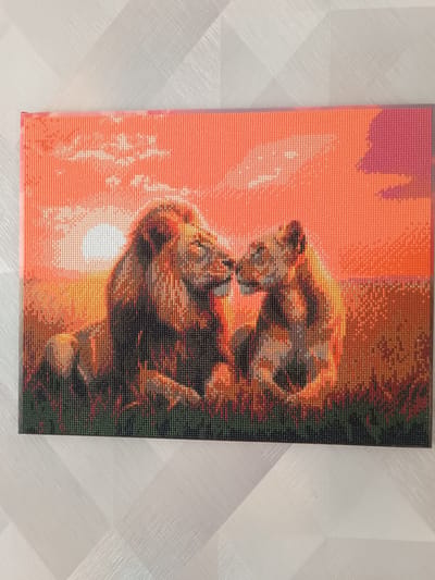 A diamond painting depicts a male and female lion nuzzling each other against a vibrant sunset. The male lion is on the left, with a full mane, while the female lion is to its right. They are seated in tall grass, with the sun a bright white orb low on the horizon behind them. The sky is a gradient of oranges and pinks, with some wispy clouds. The overall impression is one of warmth and companionship. This diamond painting is located in the city of Lisakovsk, Kazakhstan, displayed on a white, textured wall.