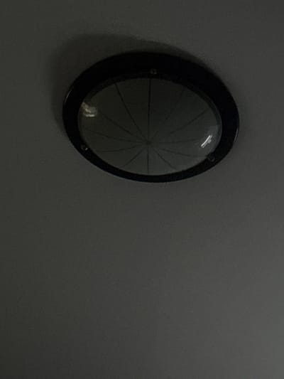The image shows a ceiling light fixture with a black frame and a clear, patterned glass cover. The light fixture is centrally located in the upper portion of the frame against a dark gray, textured ceiling. The glass cover has a radial pattern, with lines emanating from the center. There are reflections visible on the glass, suggesting an interior environment. The image is dark, with the light fixture being the primary source of illumination in the scene. There are no people or other objects visible. The image is taken from a low angle, looking up at the ceiling.