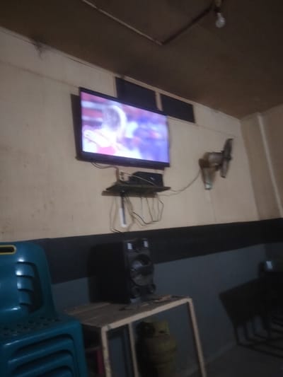 The interior scene features a wall-mounted television displaying a sporting event or show. A shelf beneath the television holds a set-top box and assorted cables. A rotary fan is mounted on the wall to the right of the television. Below, a black speaker sits on a small metal table, next to a green propane tank. A stack of teal plastic chairs is visible in the foreground to the left. The walls are beige, with a dark stripe at approximately waist height. The ceiling is low, made of what appears to be wood panels, and a bare lightbulb is visible. The location is Kruengmanyang, Indonesia.