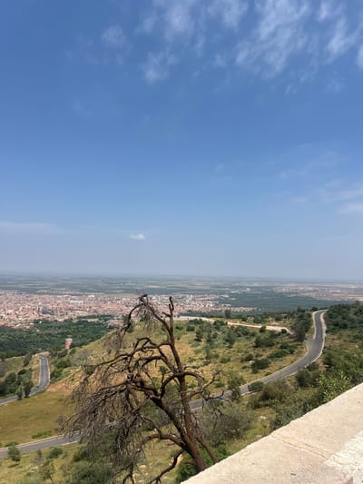 The image shows a landscape view overlooking the city of Beni Mellal, Morocco. The foreground features part of a wall structure, possibly belonging to a high point or viewing platform. A bare, branching tree is present, extending towards the cityscape. The middle ground consists of a landscape with a winding road visible on a hillside, leading towards the city. The city is spread out across the plain, showing residential buildings and some greenery. The sky occupies a significant portion of the image, transitioning from a clear blue to a hazy horizon.