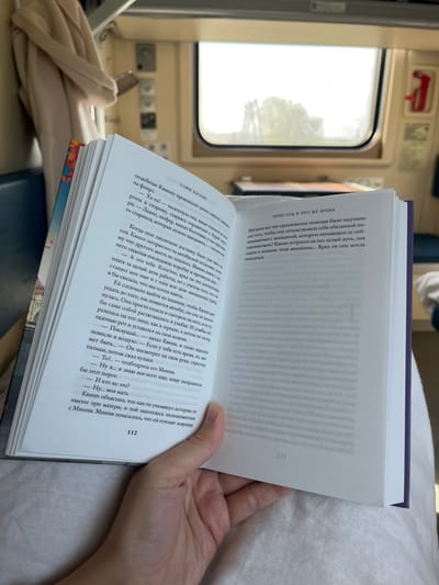 The image shows a person reading a book titled "Through the Year in the Same Time" by Софи Касенс (Sofi Kasens) in what appears to be a train compartment. The book is open to pages 112 and an unidentified page, and the person's hand is holding the open book. The window of the train is visible in the background. The environment suggests the individual is traveling.