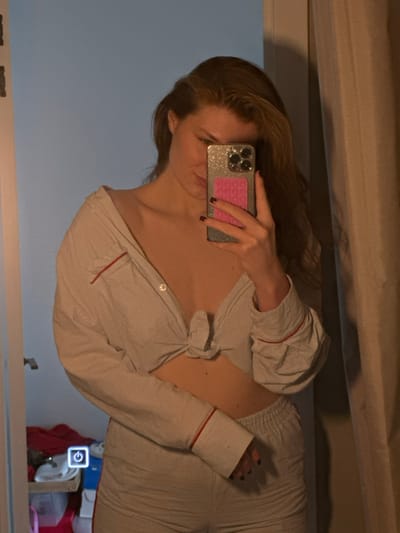 An adult female is captured in a mirror selfie indoors. She possesses reddish-brown hair and is wearing a light-colored, vertically striped two-piece outfit with red piping details; the top is a long-sleeved shirt unbuttoned and tied at the waist, exposing the midriff, and the bottom consists of matching shorts. Her visible fingernails are painted a dark hue. She holds a smartphone featuring a glittery silver case and a pink circular accessory on its back, partially obscuring her face, which exhibits a subtle smirk. The reflected environment reveals a light-colored surface on the left, a dark curtain or wall on the right, and on the lower left, a surface containing a white object, red fabric, and an illuminated rectangular display showing a power symbol. The specific location in the city of Бакча-Сарай, Russia, cannot be identified from the visual content of the image.