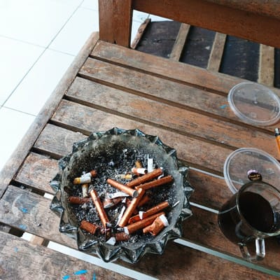 The image shows a wooden table top with an ashtray in the center, filled with cigarette butts. The ashtray is made of dark grey ceramic with a starburst pattern. There is a glass of black coffee in the bottom right corner of the image. There is a plastic container on the right side of the ashtray with a white sticker that reads "Oven Safe." To the right of the container is a brown wooden pencil. The table is made of wood with a reddish-brown stain and has a worn, weathered appearance. The coordinates provided locate the scene to be in Pameungpeuk, Indonesia.  The image suggests a moment of relaxation and casualness, with evidence of cigarette smoking.  The scene seems to be taking place outdoors.