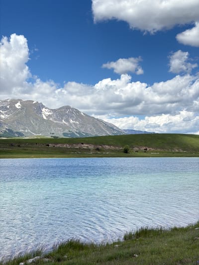 This image depicts a natural landscape in Javorje, Montenegro, under a partly cloudy sky during daylight hours. The upper portion of the frame features a vibrant blue sky populated by numerous white cumulus clouds of varying sizes and densities. In the background, a range of large, rugged mountains rises, exhibiting grey and green rock formations with visible patches of snow on their peaks and upper slopes. Below the mountains, a broad band of lush green hills stretches across the midground, appearing to be covered predominantly by grass. A distinct line of denser, darker green vegetation, possibly trees, is visible along the base of the mountains. The central and lower-mid sections of the image are dominated by a tranquil body of water, likely a lake, which displays a gradient of blue and turquoise hues, indicating clarity and potentially varying depths. The surface of the water shows subtle ripples. The immediate foreground consists of a grassy bank, green and relatively short, extending down to the water's edge. No human figures or discernible man-made structures are visible within the frame.